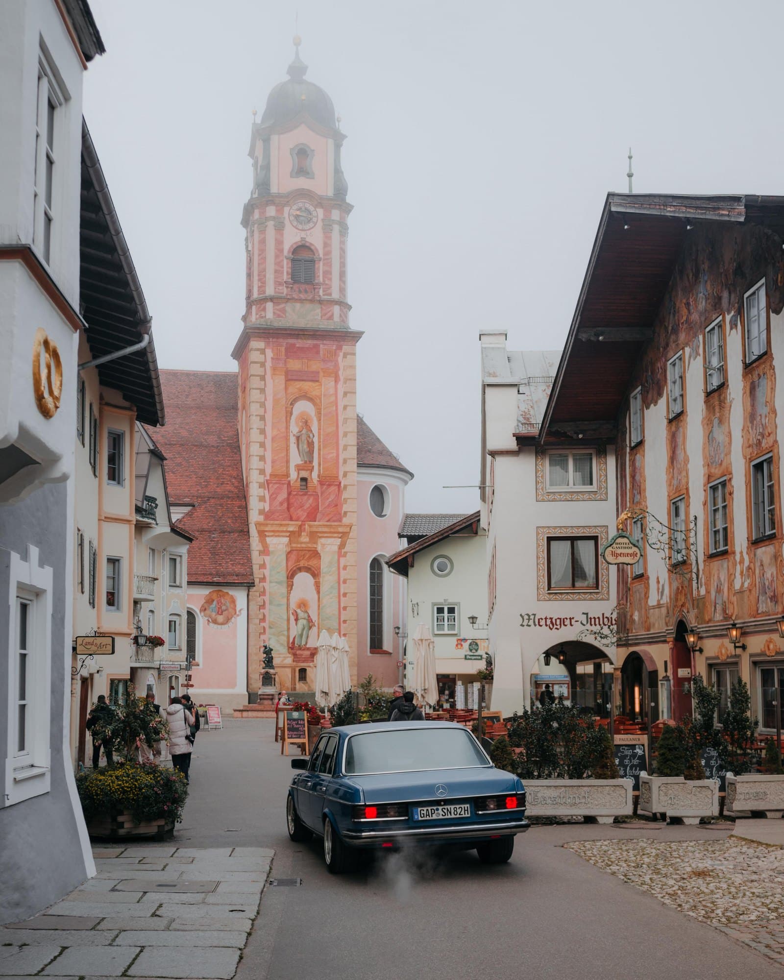 An old Mercedes saloon driving away down a Bavarian street of painted houses, towards a church tower in the morning haze.